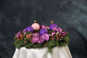 A photo of four round candles placed on a pine wreath with decorative globes, pinecones, and ribbons. Three of the candles are violet and one of them is rose. The rose candle and two of the purple candles are lit, signifying the third Sunday of Advent.
