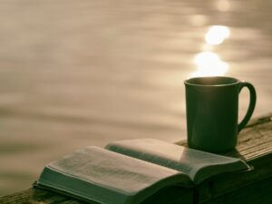 A photo of an open Bible and a coffee mug on a wooden railing with the sun reflecting off of water in the background