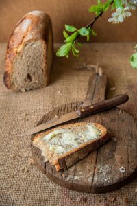 A buttered piece of bread sits next to a knife on a cutting board. In the background is a loaf of bread, with a plant hanging from above.