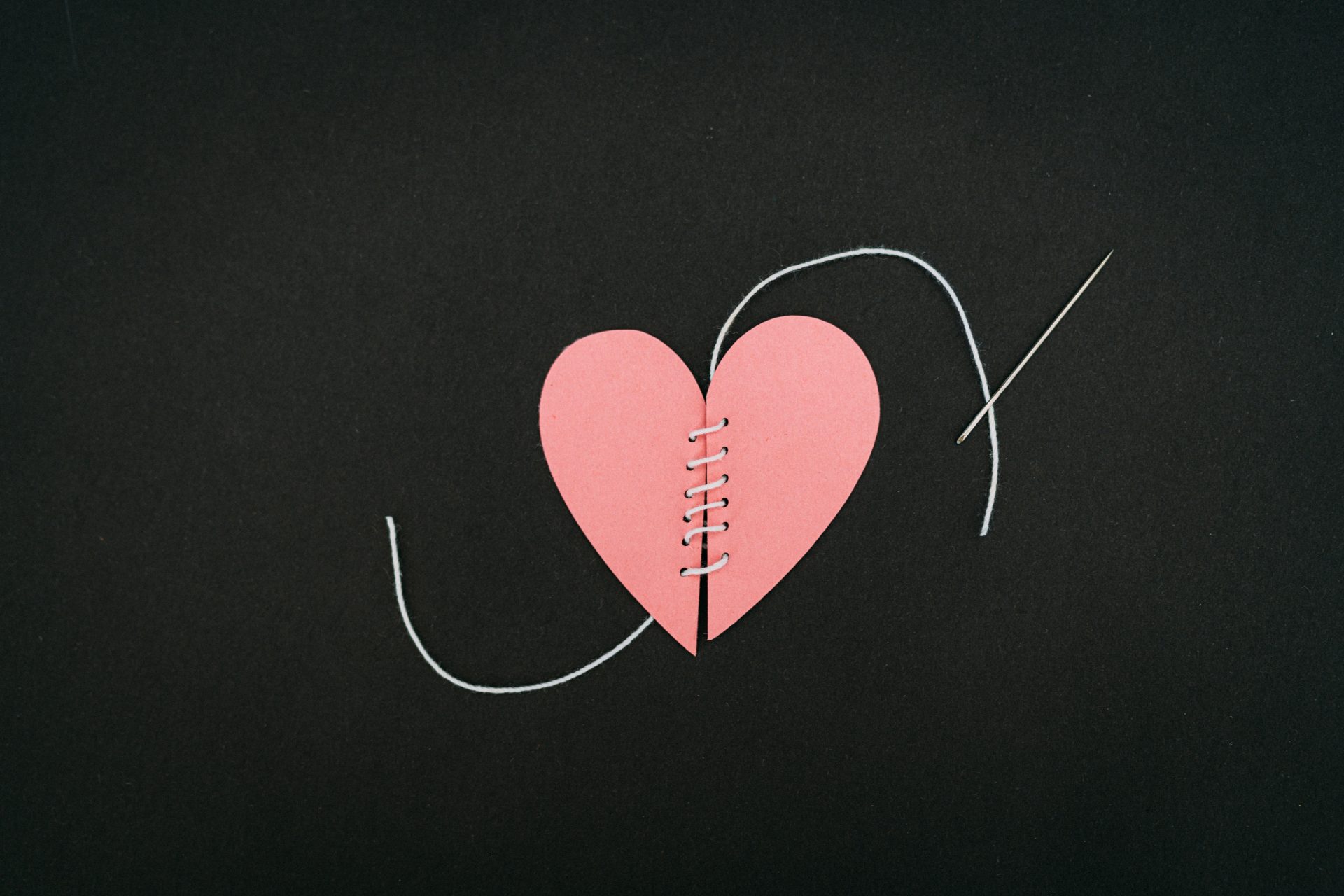 Broken heart sewn back together (kelly-sikkema-wC2HUqVV2Go-unsplash) A photo of a red paper heart cut in two, with sewn thread bringing the two halves back together against a black background, with a sewing needle laying nearby