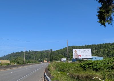 A mid-range photo of the 2026 billboard put up by Sacred Heart Radio, featuring an image of a priest holding up a monstrance, with the message "We love our priests. God bless, we're praying for you" from Sacred Heart Radio