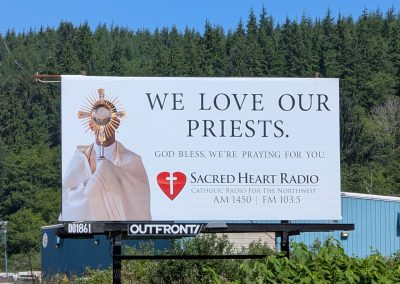 A close-up photo of the 2026 billboard put up by Sacred Heart Radio, featuring an image of a priest holding up a monstrance, with the message "We love our priests. God bless, we're praying for you" from Sacred Heart Radio