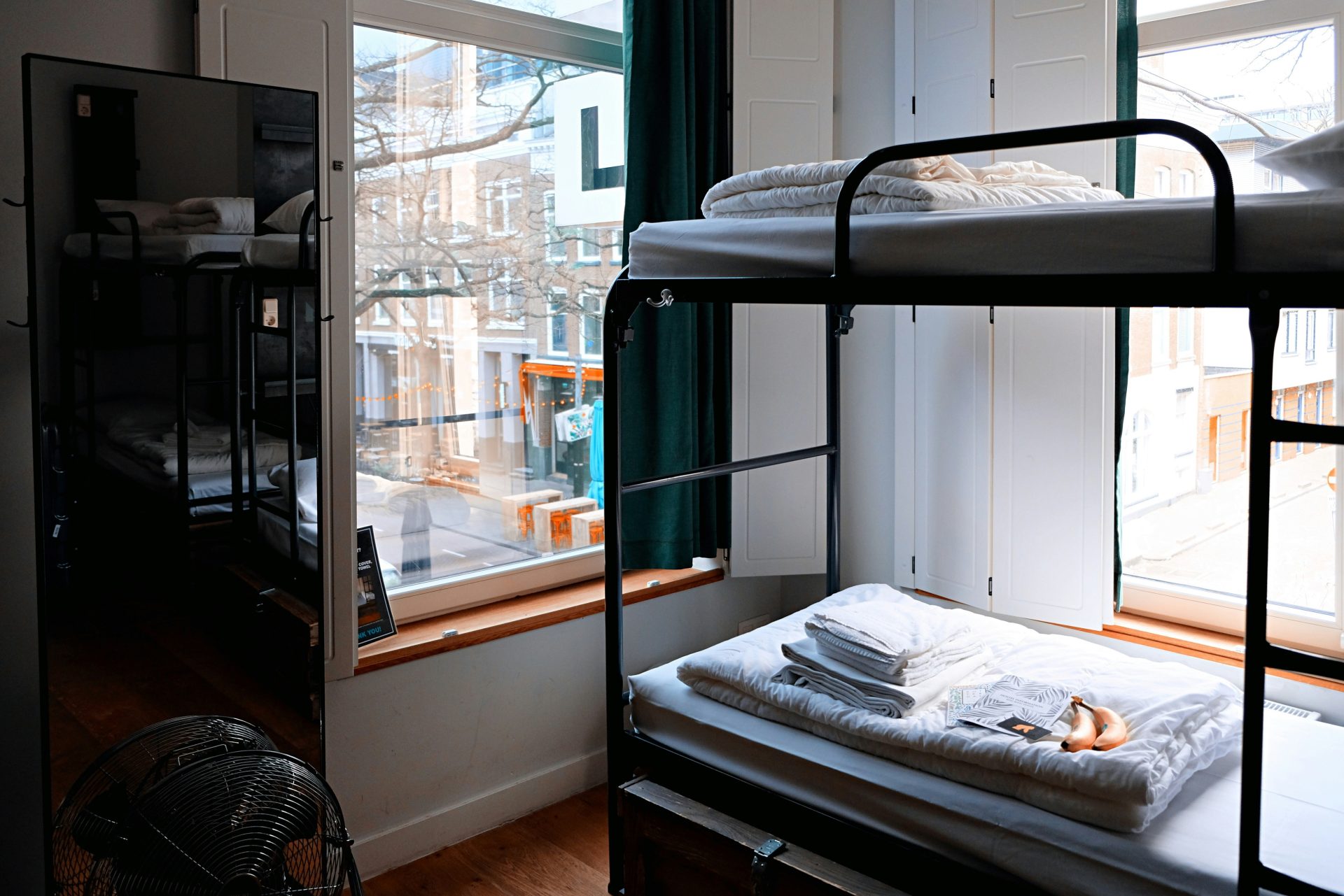 marcus-loke-WQJvWU_HZFo-unsplash A metal frame dormitory bunk bed in the corner of an dorm, in front of two windows. On each bed are folded sheets and pillow cases