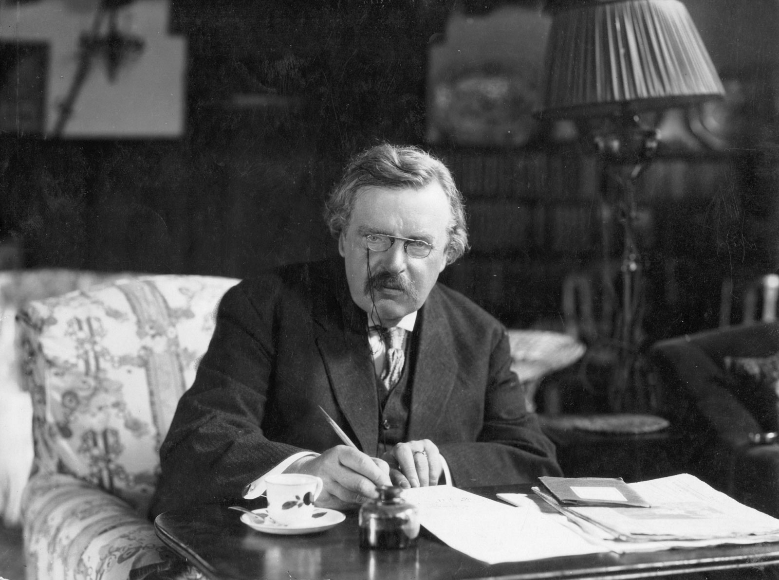 A photograph of G.K. Chesterton in an armchair at his desk, pen in hand. Wearing a three-piece suit and glasses, he looks at the camera.